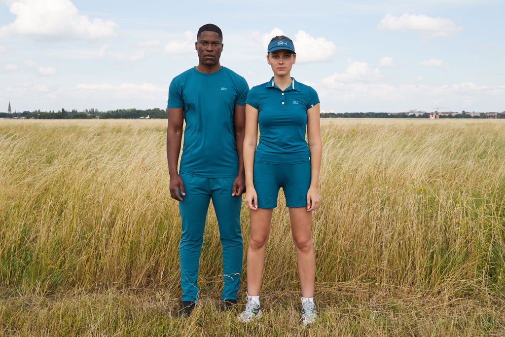 Frau mit petrolfarbener Sportkleidung bestehend aus Polo Shirt, Shorts und Sun Visor und Mann mit petrolfarbenen Shirt und langer Hose